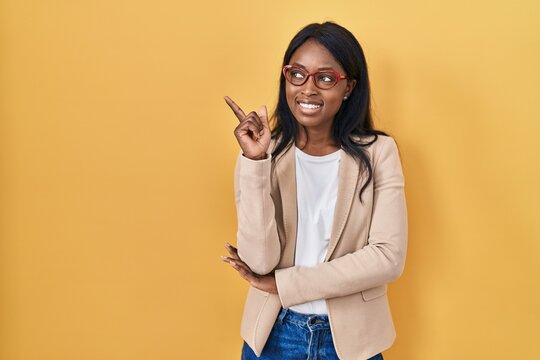 African young woman wearing glasses with a big smile on face, pointing with hand and finger to the side looking at the camera.