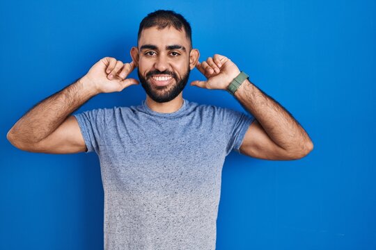 Middle east man with beard standing over blue background smiling pulling ears with fingers, funny gesture. audition problem