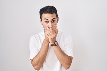 Handsome hispanic man standing over white background shocked covering mouth with hands for mistake....