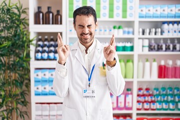 Handsome hispanic man working at pharmacy drugstore gesturing finger crossed smiling with hope and eyes closed. luck and superstitious concept.