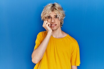 Young modern man standing over blue background looking stressed and nervous with hands on mouth biting nails. anxiety problem.