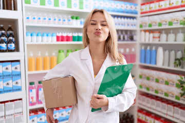 Young caucasian woman working at pharmacy drugstore holding box doing inventory looking at the camera blowing a kiss being lovely and sexy. love expression.