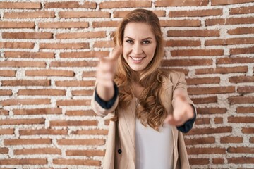 Beautiful blonde woman standing over bricks wall looking at the camera smiling with open arms for hug. cheerful expression embracing happiness.