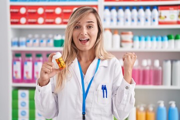 Young blonde woman working at pharmacy drugstore holding pills screaming proud, celebrating victory and success very excited with raised arms