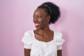 African woman with curly hair standing over pink background looking away to side with smile on face, natural expression. laughing confident.