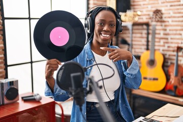 Beautiful black woman holding vinyl record at music studio smiling happy pointing with hand and finger