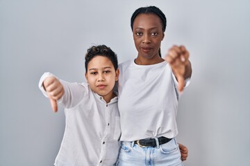 Young mother and son standing together over white background looking unhappy and angry showing rejection and negative with thumbs down gesture. bad expression.