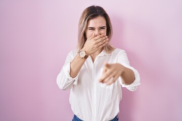 Young beautiful woman standing over pink background laughing at you, pointing finger to the camera with hand over mouth, shame expression