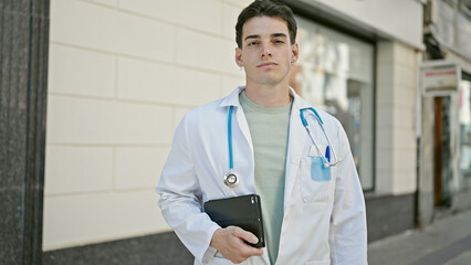 Young hispanic man doctor standing with serious expression holding touchpad at hospital