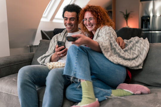 Interracial couple sitting on couch, snuggled in blankets, eating popcorn and scrolling through movies on television while talking