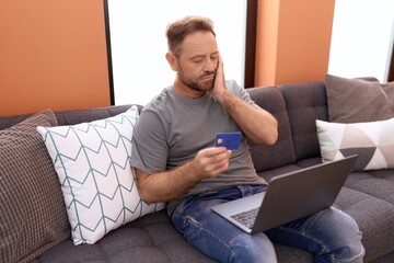 Middle age man using laptop and credit card sitting on sofa at home