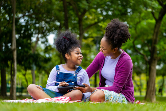 African American mother is teaching her young daughter to read using digital tablet while having a summer picnic in the public park for education and happiness concept