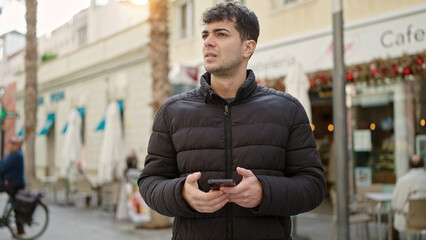 Young hispanic man using smartphone looking away at coffee shop terrace