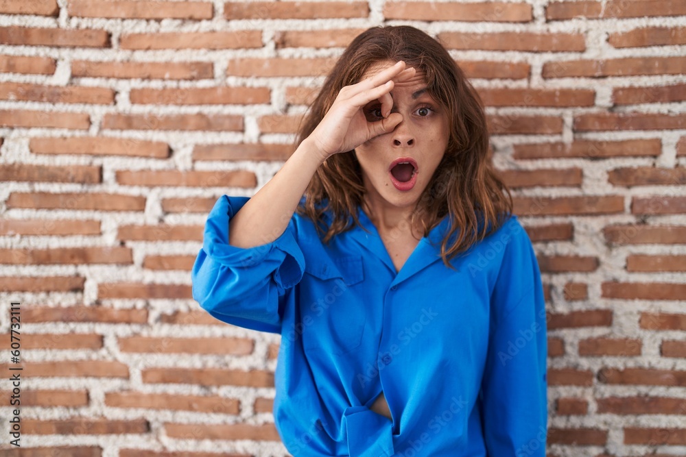 Sticker Beautiful brunette woman standing over bricks wall doing ok gesture shocked with surprised face, eye looking through fingers. unbelieving expression.