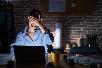 Beautiful brunette woman working at the office at night smiling and laughing with hand on face covering eyes for surprise. blind concept.