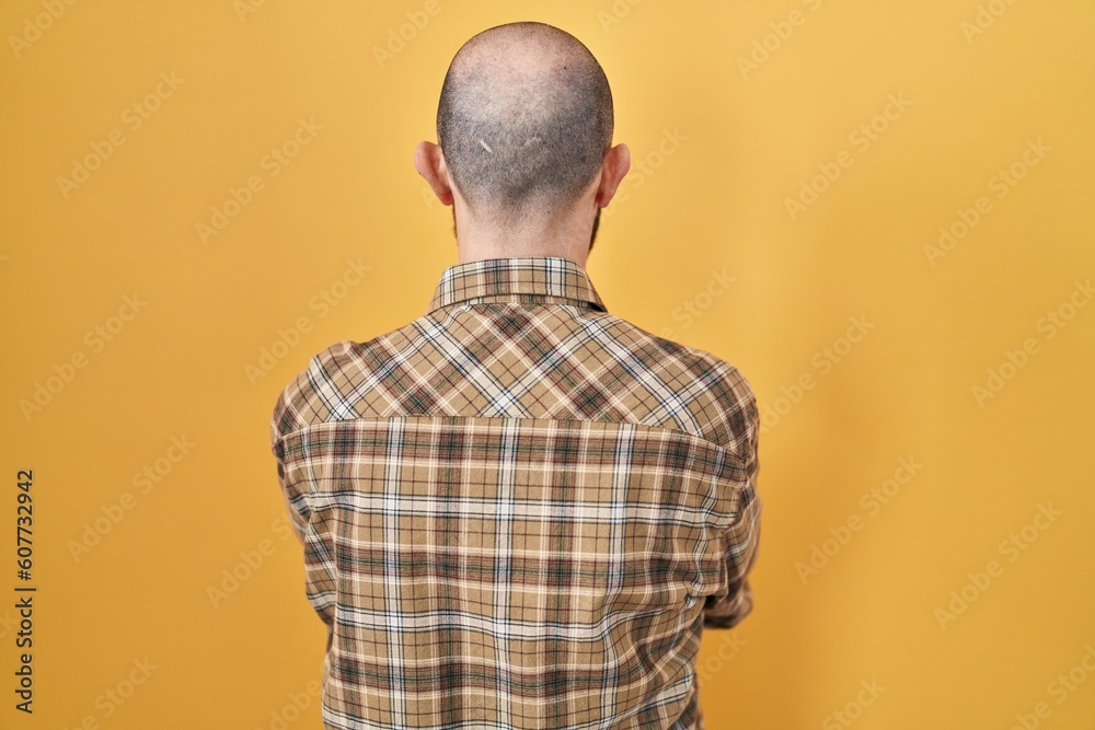 Poster young hispanic man with tattoos standing over yellow background standing backwards looking away with