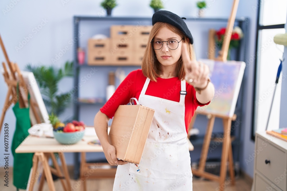 Poster Young redhead woman at art studio holding art case pointing with finger up and angry expression, showing no gesture