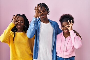 Group of three young black people standing together over pink background doing ok gesture with hand smiling, eye looking through fingers with happy face.
