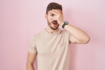 Hispanic man with beard standing over pink background peeking in shock covering face and eyes with...