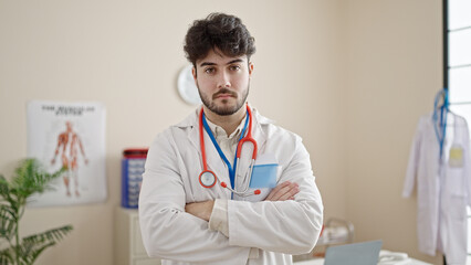 Young hispanic man doctor standing with serious expression and arms crossed gesture at clinic