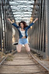 Fototapeta na wymiar smiling red-haired girl dressed in urban clothes jumping on old railway tracks of a steel girder bridge