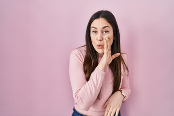 Young brunette woman standing over pink background hand on mouth telling secret rumor, whispering malicious talk conversation