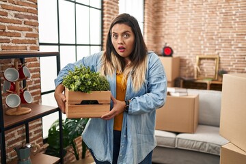 Young hispanic woman moving to a new home holding plants scared and amazed with open mouth for...