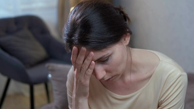 A sad Caucasian brunette woman lowers her head and wearily touches her forehead with her hand while in a depressed state, worrying about bad news. Mental health and fatigue