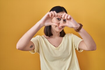 Hispanic girl wearing casual t shirt over yellow background doing heart shape with hand and fingers smiling looking through sign