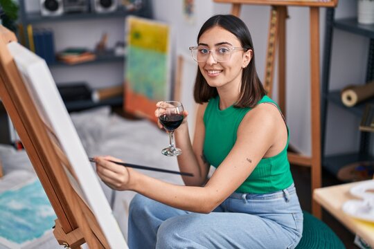 Young hispanic woman artist drinking wine drawing at art studio
