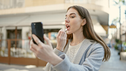 Young hispanic woman using smartphone as a mirror applying lipstick at coffee shop terrace