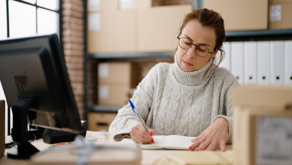 Middle age hispanic woman ecommerce business worker writing on book at storehouse office