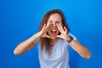 Fototapeta premium Brunette woman standing over blue background shouting angry out loud with hands over mouth