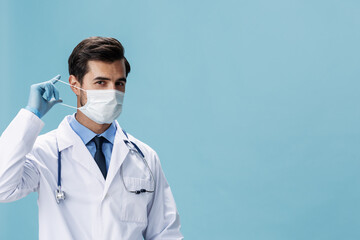 Male doctor in a white coat and medical mask looks at the camera on a blue isolated background, copy space, space for text