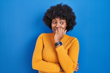 Fototapeta na wymiar Black woman with curly hair standing over blue background looking stressed and nervous with hands on mouth biting nails. anxiety problem.