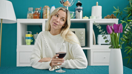 Young blonde woman drinking glass of wine sitting on table speaking at dinning room