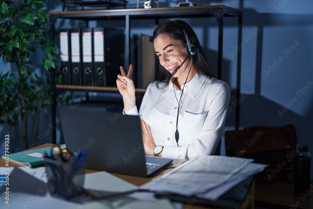 Wall mural Young brunette woman wearing call center agent headset working late at night smiling with happy face winking at the camera doing victory sign. number two.