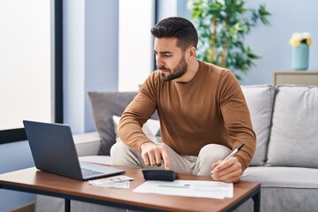 Naklejka premium Young hispanic man accounting economy sitting on sofa at home