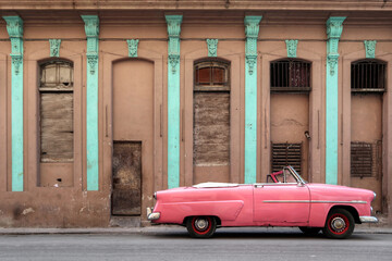 A side photo of a pink car in Havana, Cuba park in front of an brown facade 