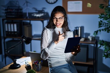 Young brazilian woman using touchpad at night working at the office pointing fingers to camera with happy and funny face. good energy and vibes.