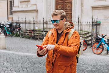 Fototapeta premium Cheerful young guy walking on city street with modern smartphone in hands. Handsome man in sunglasses reading some good and smiling. Technology for lifestyles.