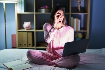 Young hispanic woman using computer laptop on the bed doing ok gesture shocked with surprised face, eye looking through fingers. unbelieving expression.