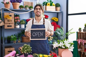 Hispanic man with long hair working at florist holding open sign sticking tongue out happy with...