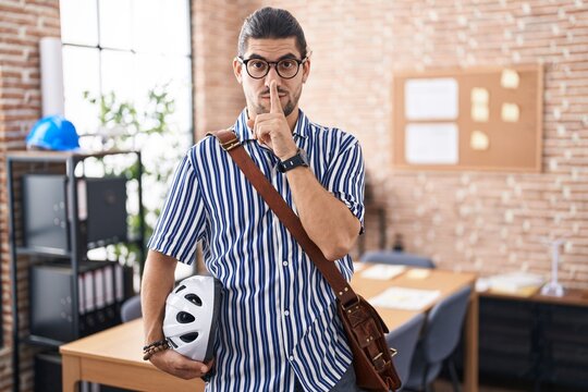 Hispanic man with long hair working at the office holding bike helmet asking to be quiet with finger on lips. silence and secret concept.