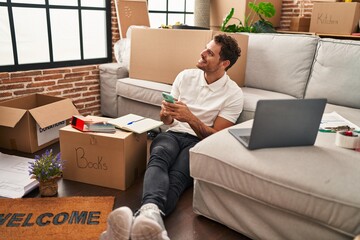 Young hispanic man using smartphone sitting on floor at new home