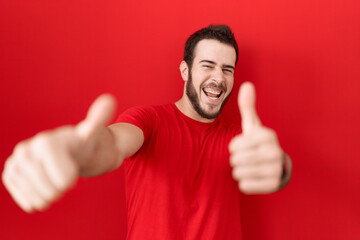 Young hispanic man wearing casual red t shirt approving doing positive gesture with hand, thumbs up smiling and happy for success. winner gesture.