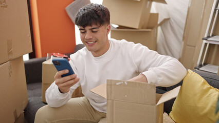 Young hispanic man unpacking cardboard box using smartphone at new home