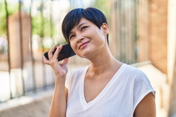 Middle age chinese woman smiling confident listening audio message by the smartphone at street