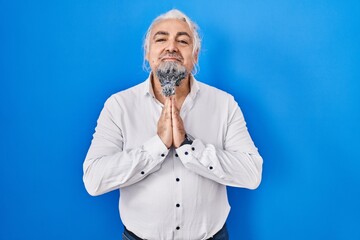 Middle age man with grey hair standing over blue background praying with hands together asking for forgiveness smiling confident.