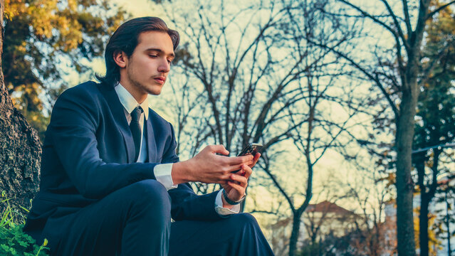 Confident man in a business suit sits outdoors and using a smartphone. Portrait of the young man in a business suit with mobile phone, in the park. Serious businessman in city looking to a cell phone.